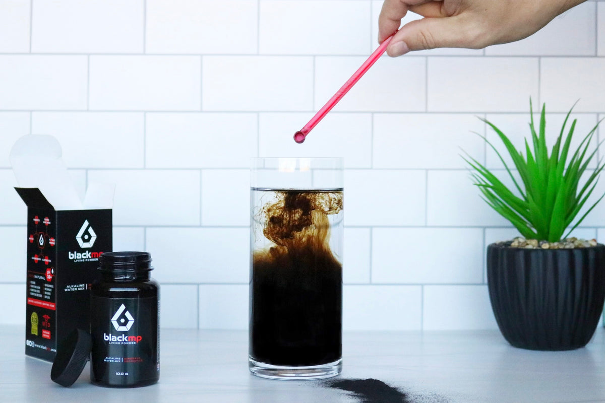 Hand scooping BlackMP Fulvic and Humic Mineral Powder into water, creating a dark alkaline drink with a red scoop.