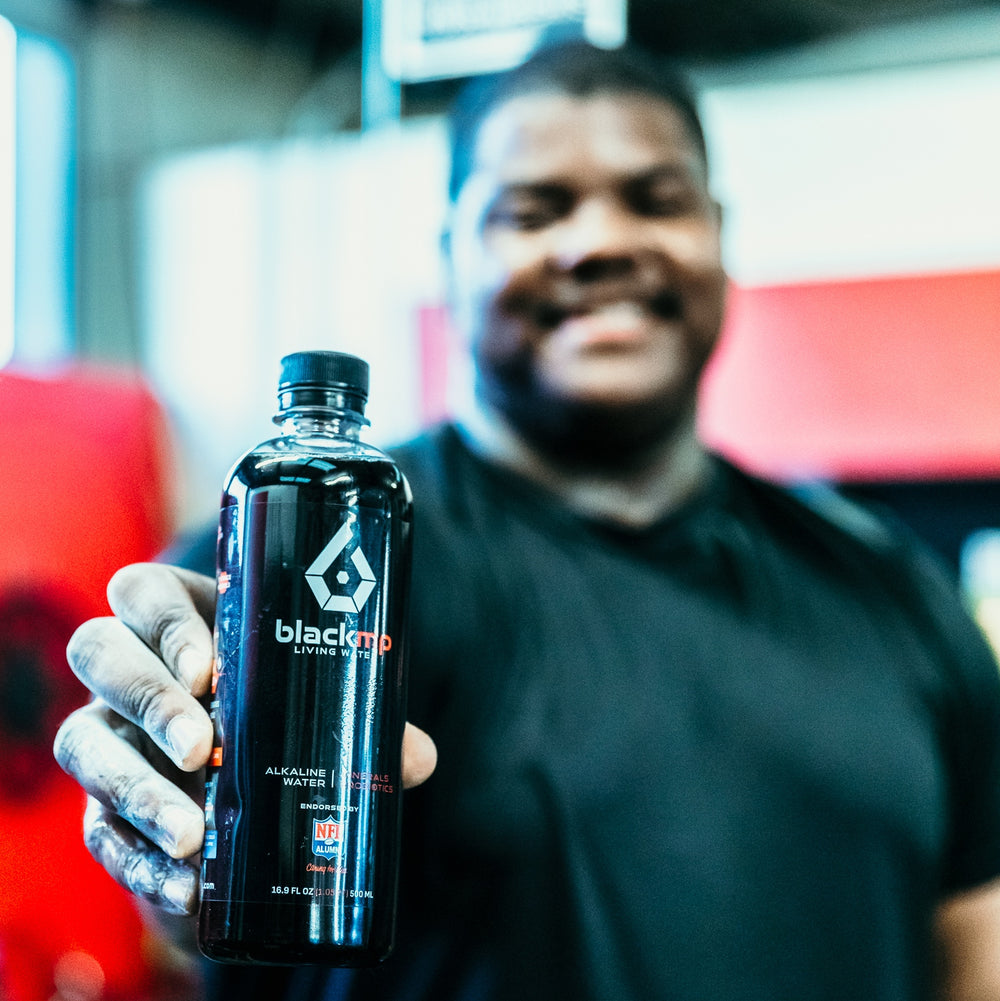 A person holding BlackMP Living Water, an alkaline water with fulvic and humic minerals, featuring an NFL Alumni logo.